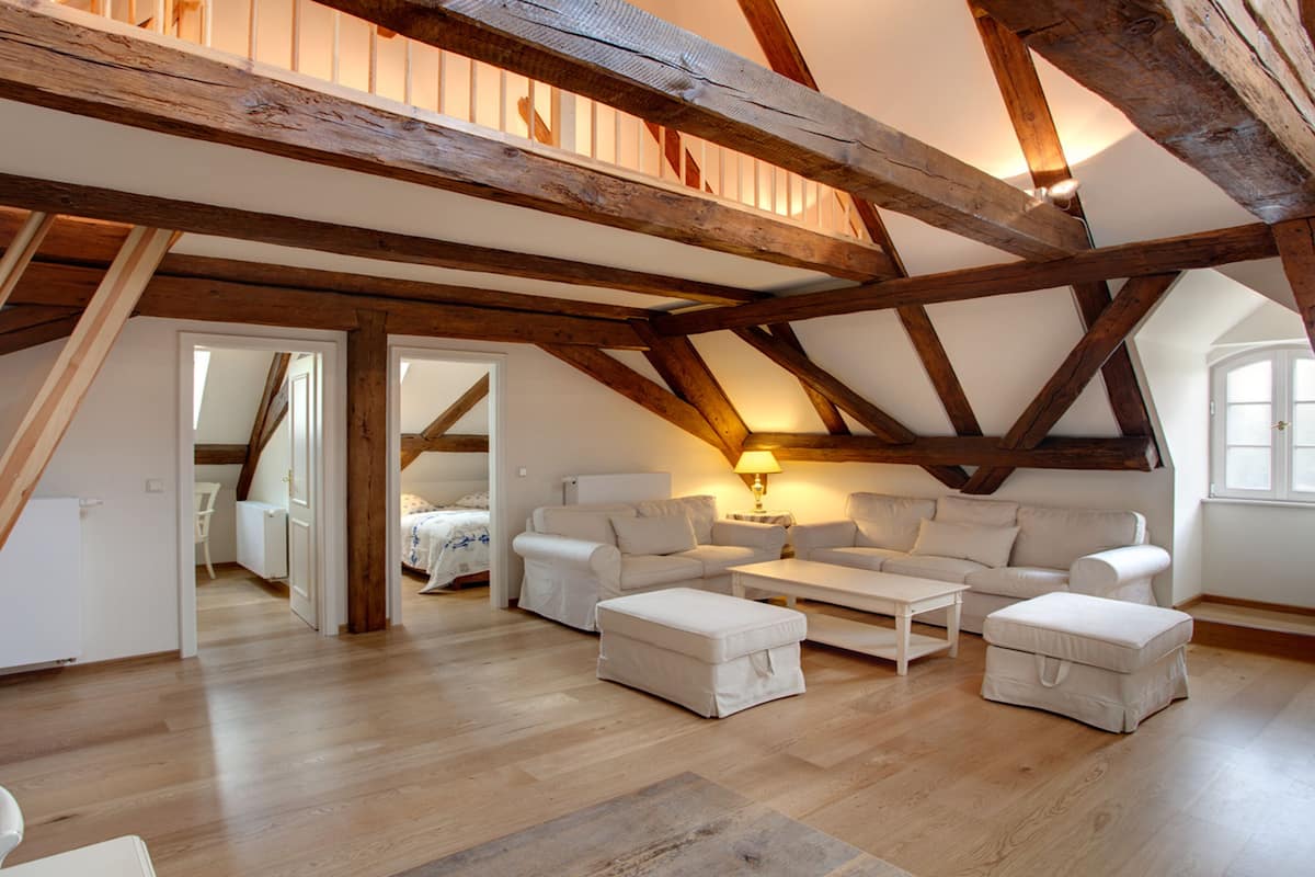 Amplio dormitorio tipo loft con techo alto, vigas de madera a la vista, un ventanal grande y una zona de estar acogedora con sofá y reposapiés.