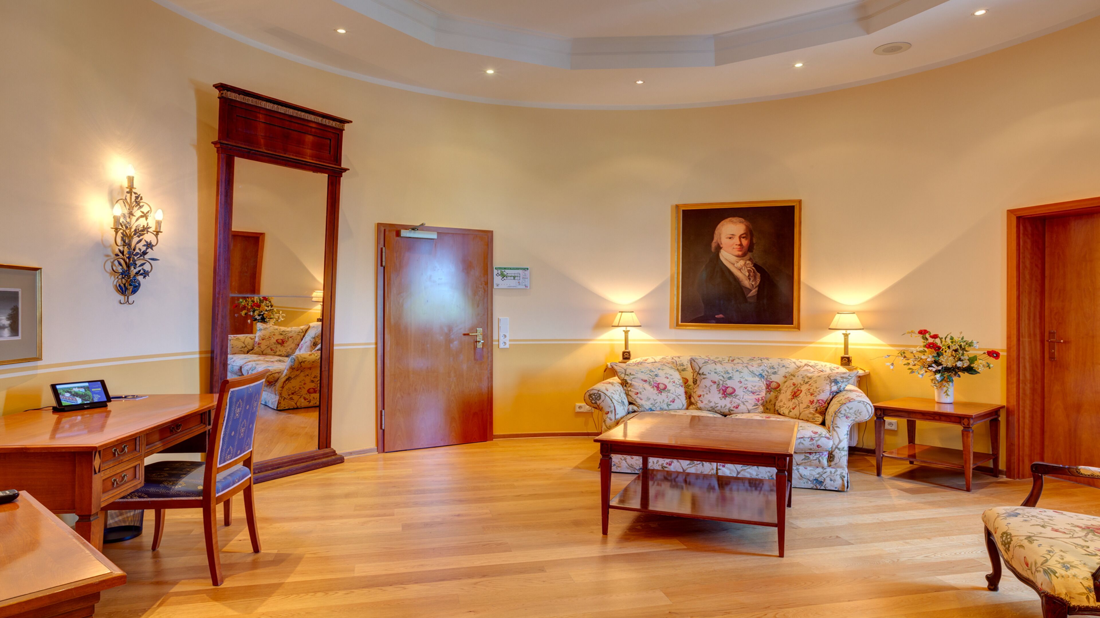 A spacious living room with a wooden floor, a floral sofa, a wooden coffee table, a desk with a laptop, and a large portrait on the wall.