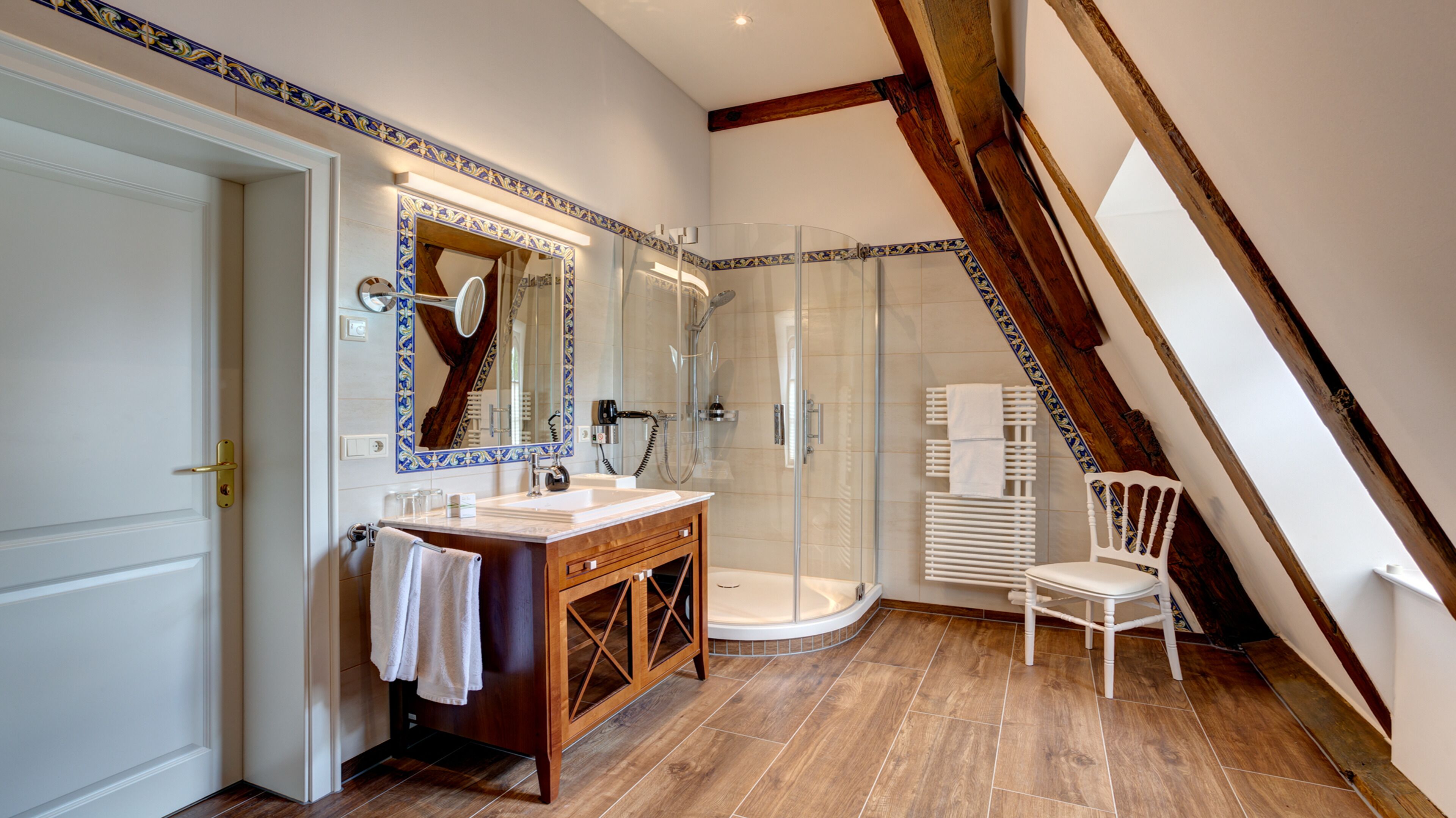 A modern bathroom with a wooden vanity, a glass shower enclosure, and a white chair.