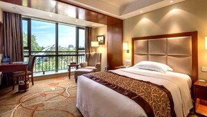 A hotel room with a large bed, a desk, a chair, and a view of the city through large windows.