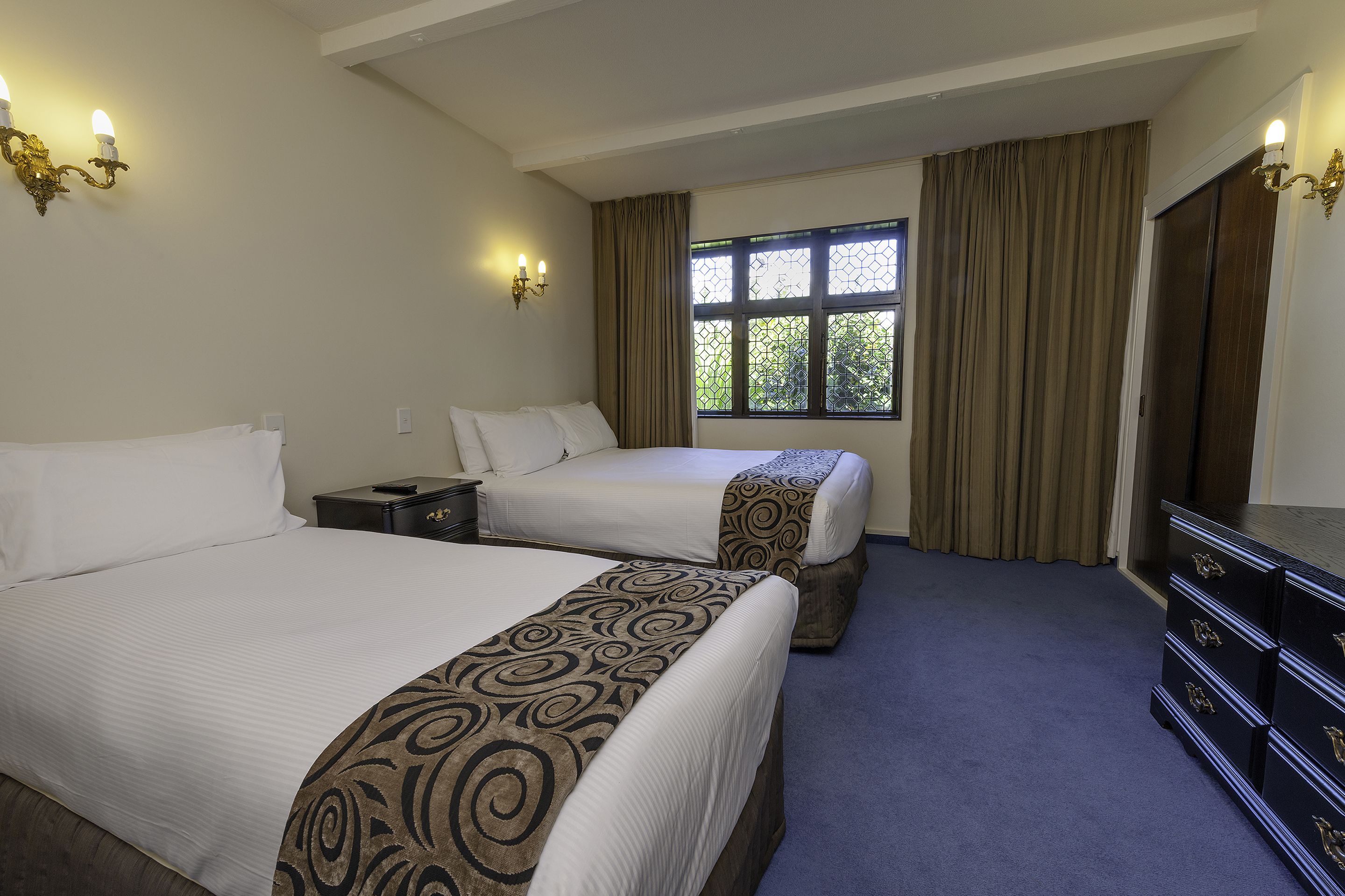 A hotel room with two beds, a window with curtains, a dresser, and wall sconces.
