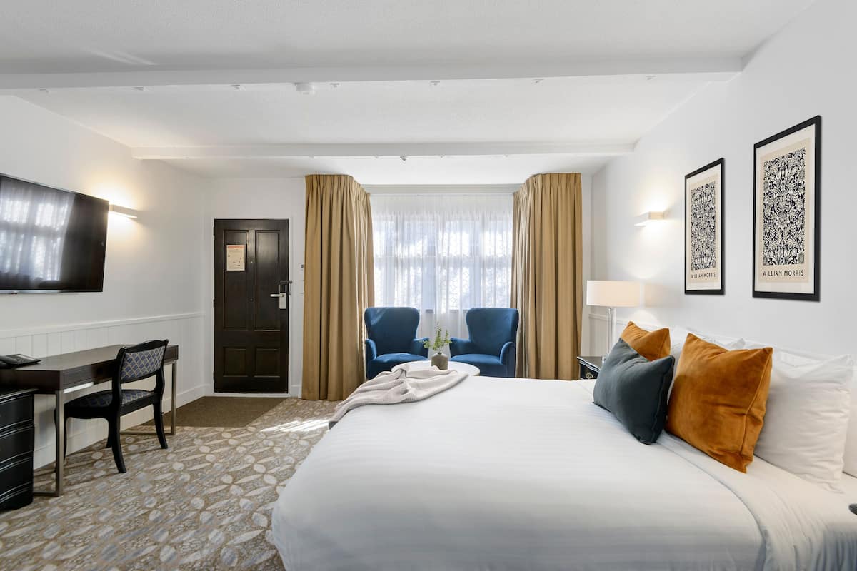 Habitación de hotel con una cama grande, un escritorio con silla, dos sillones, un televisor y cuadros en la pared.