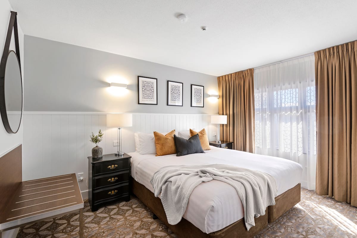 Habitación de hotel con una cama grande, una mesita de noche con lámpara, cómoda y ventana con cortinas.