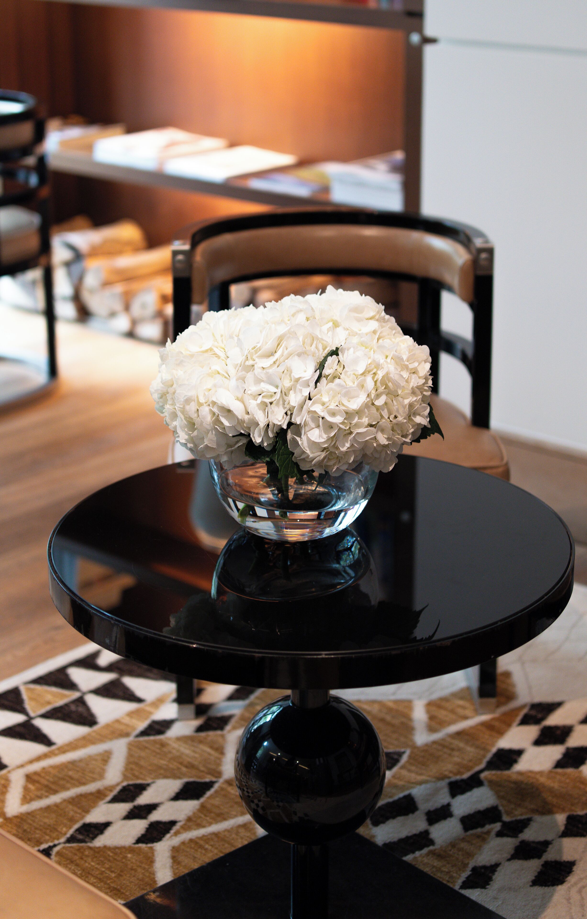 Een eettafel met een glazen vaas gevuld met witte bloemen, een zwarte tafelvoet en een kamerbreed tapijt met een patroon.