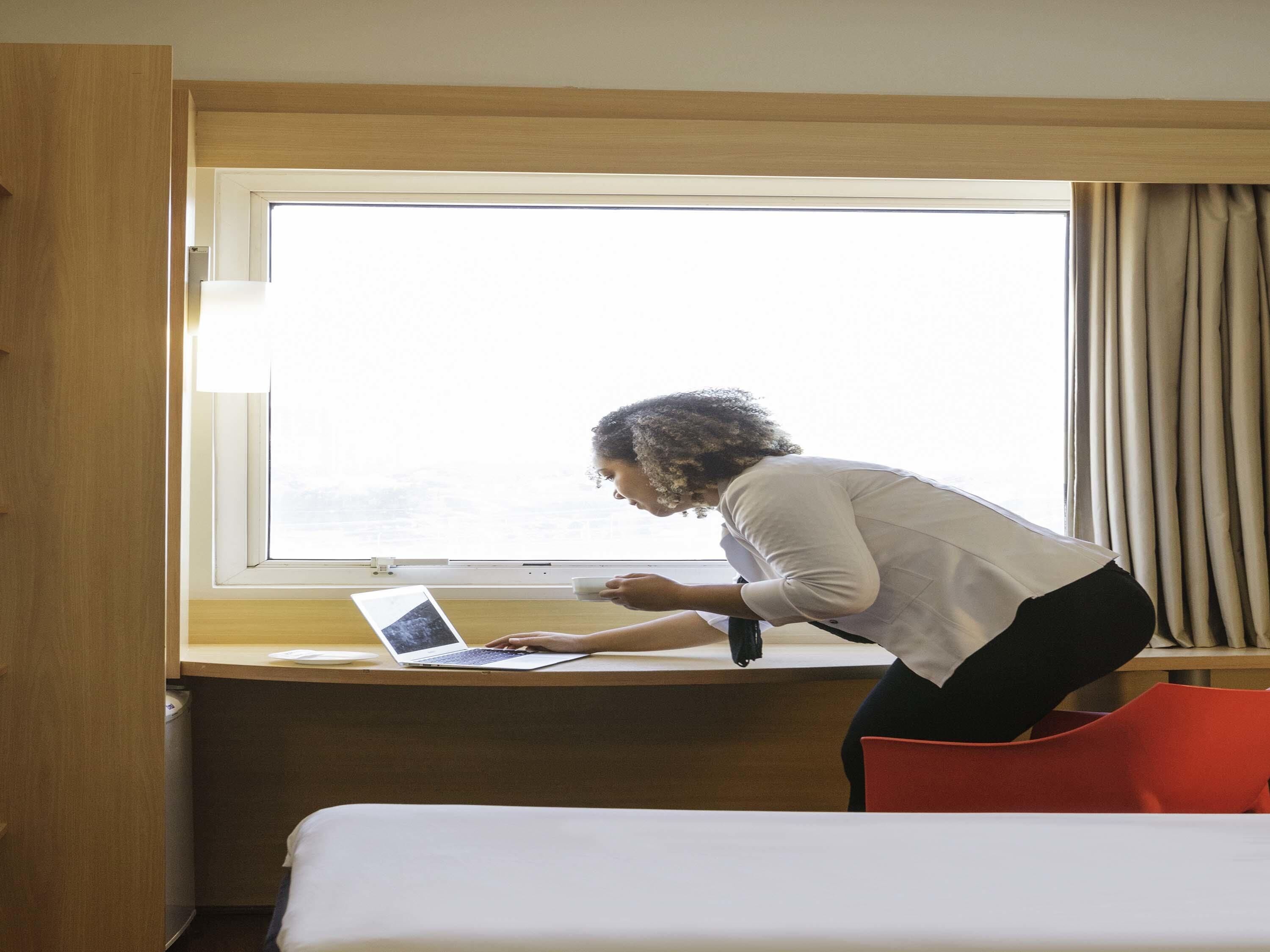 Una habitación con una cama, un escritorio con una computadora portátil, una ventana y una silla roja.