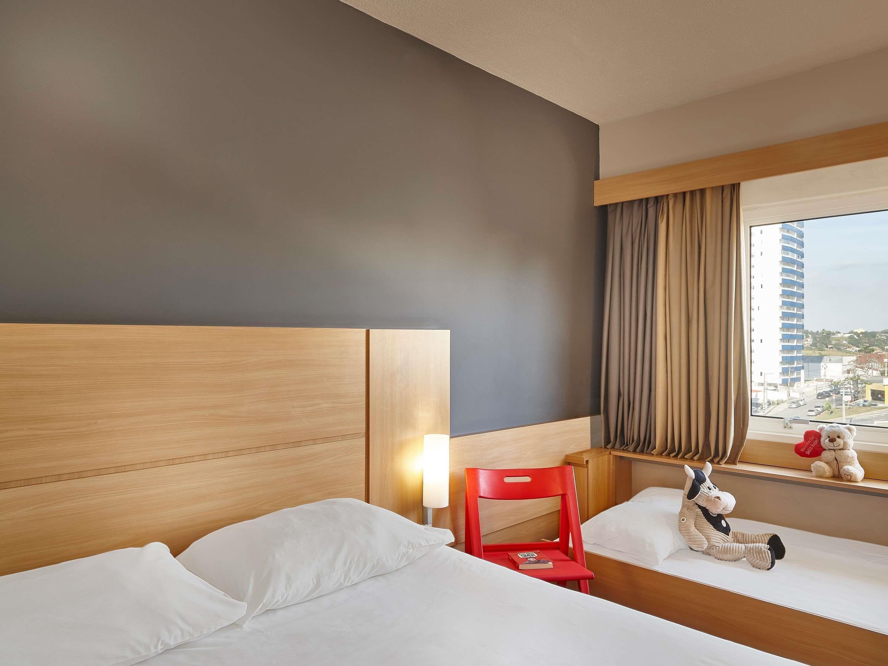 A hotel room with a wooden headboard, a red chair, a window with a view of buildings, and a stuffed toy on the windowsill.