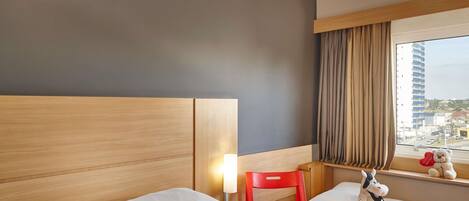 A hotel room with a wooden headboard, a red chair, a window with a view of buildings, and a stuffed toy on the windowsill.