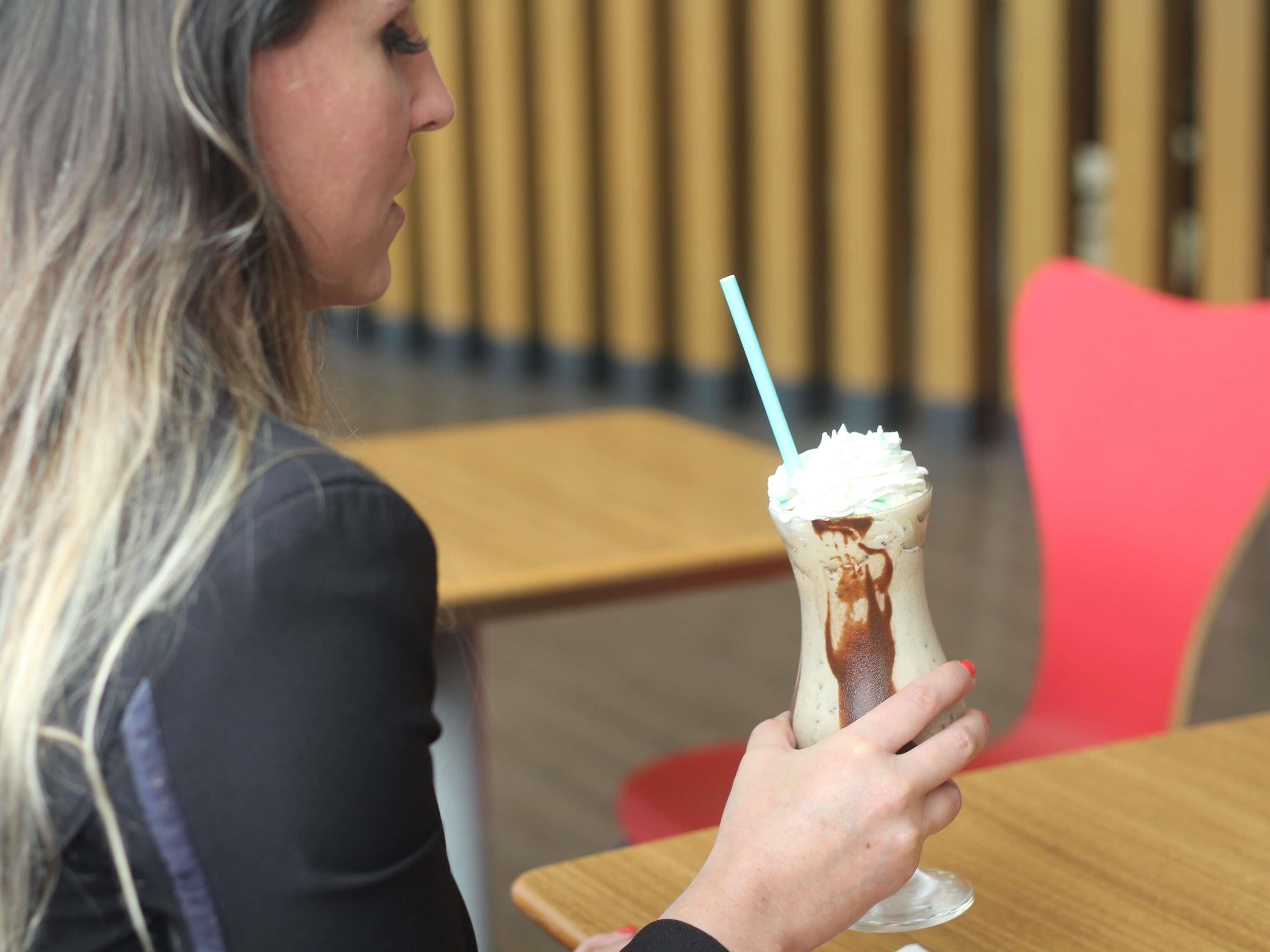 Una persona sosteniendo una bebida con crema batida y jarabe de chocolate, sentada en una mesa de madera en un café.