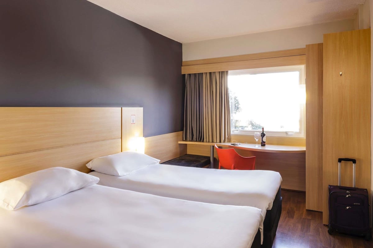 A hotel room with two beds, a desk, a chair, a window with curtains, and a suitcase.