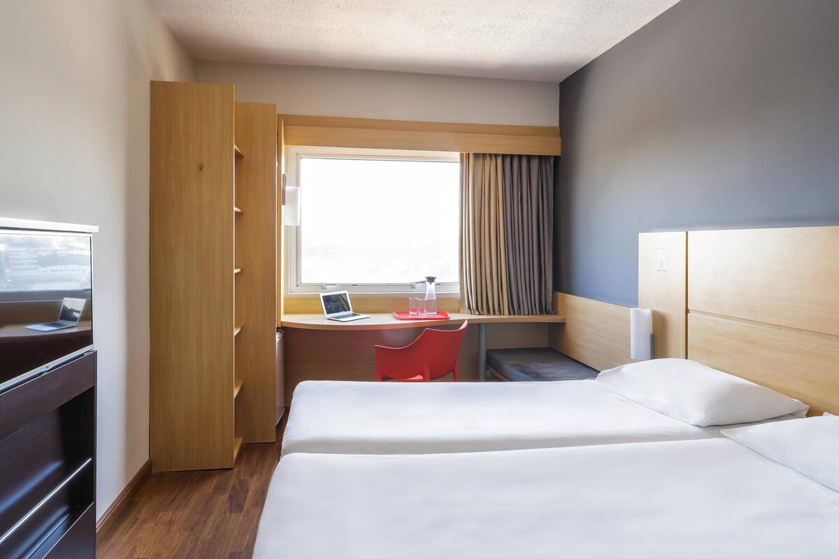 A hotel room with a bed, a desk with a laptop, a chair, and a window with curtains.