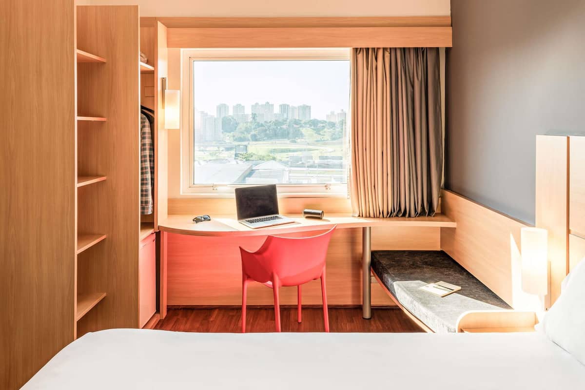 A hotel room with a desk, a chair, a bed, a window with a view of buildings, and a curtain.