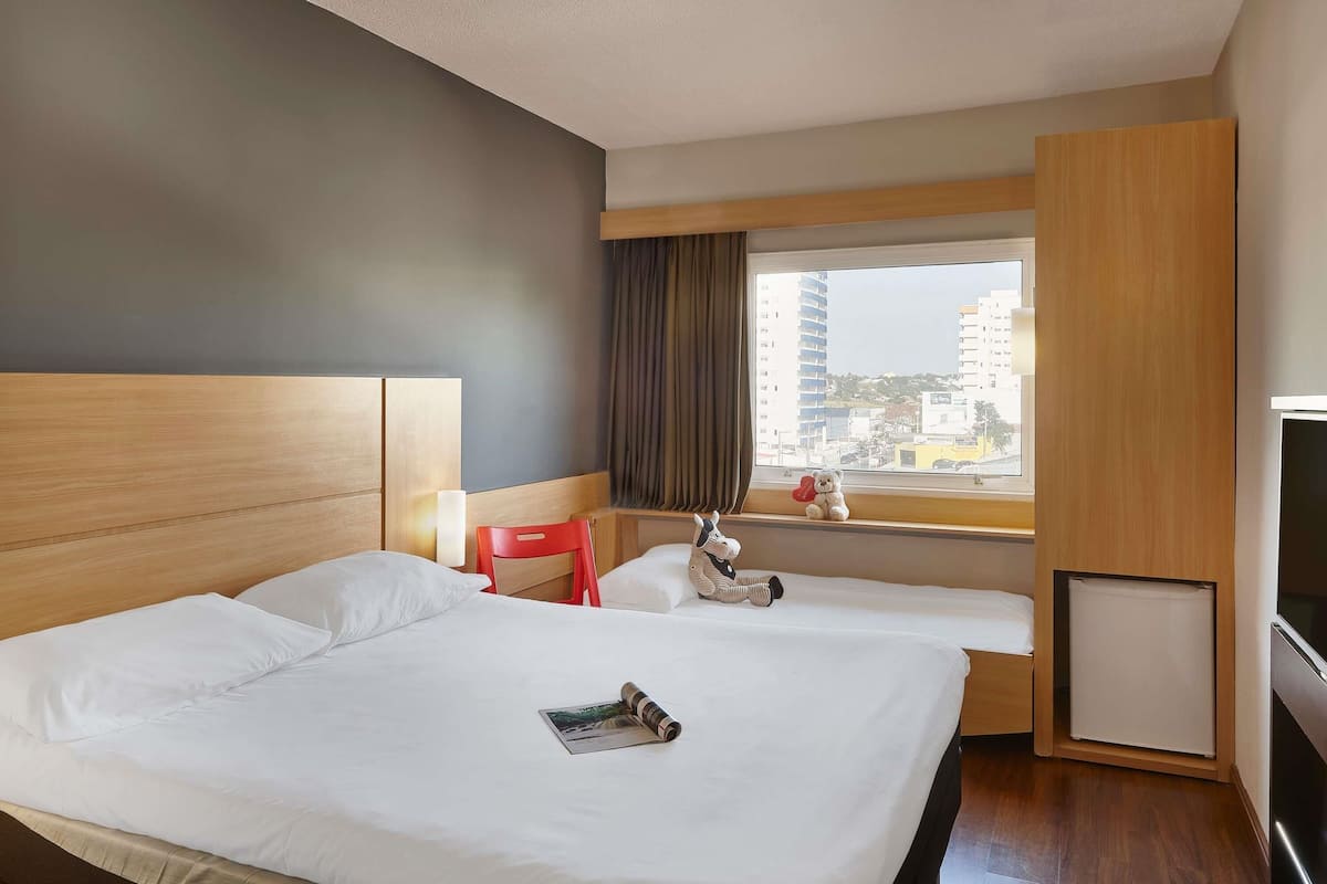 A hotel room with a large bed, a TV, a window with a view of buildings, and a red chair.
