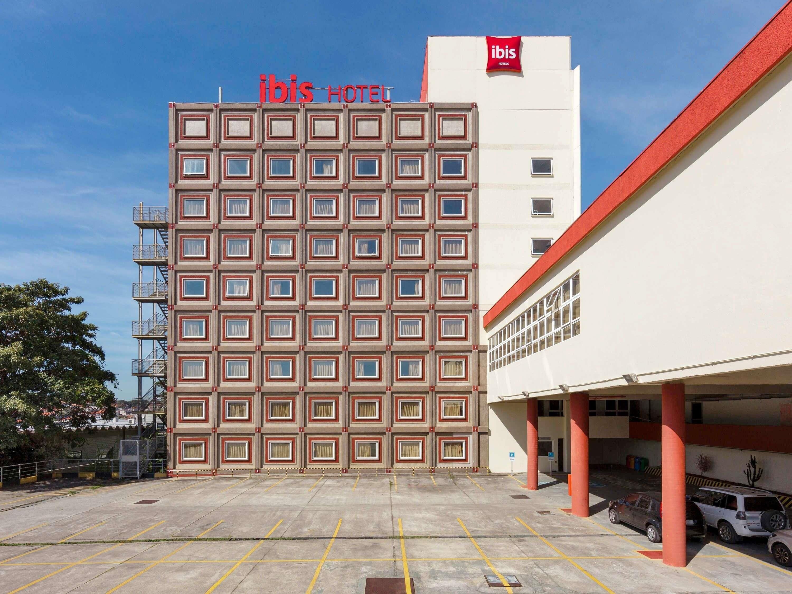 Um hotel com fachada moderna, vários andares e estacionamento com diversos carros.