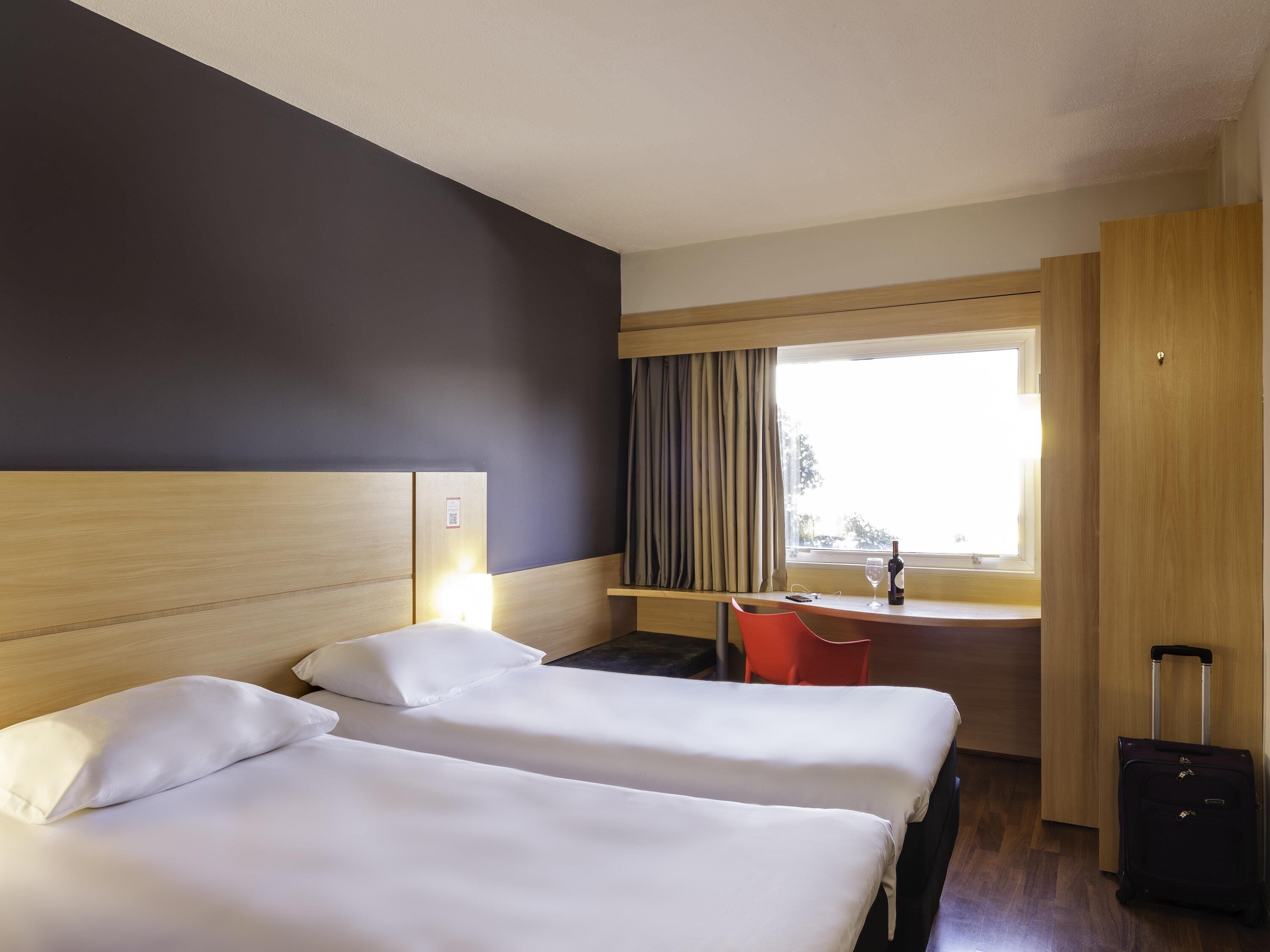 Ein Hotelzimmer mit zwei Betten, einem Schreibtisch, einem Stuhl, einem Fenster mit Vorhängen und einem Koffer.