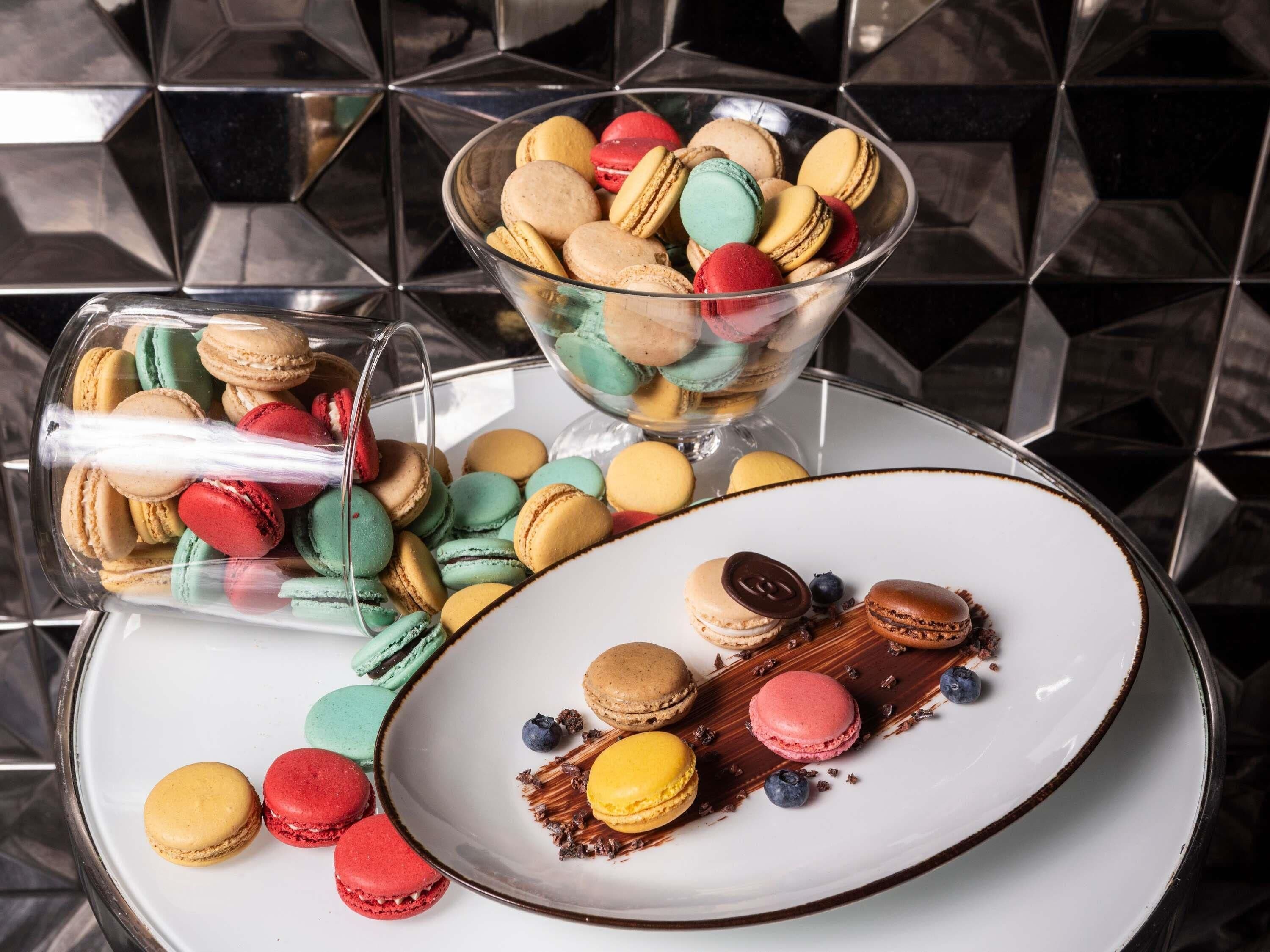 Uma tigela de vidro cheia de macarons coloridos, um prato com macarons e chocolate, e um recipiente de vidro com macarons.
