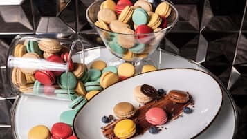 Uma tigela de vidro cheia de macarons coloridos, um prato com macarons e chocolate, e um recipiente de vidro com macarons.