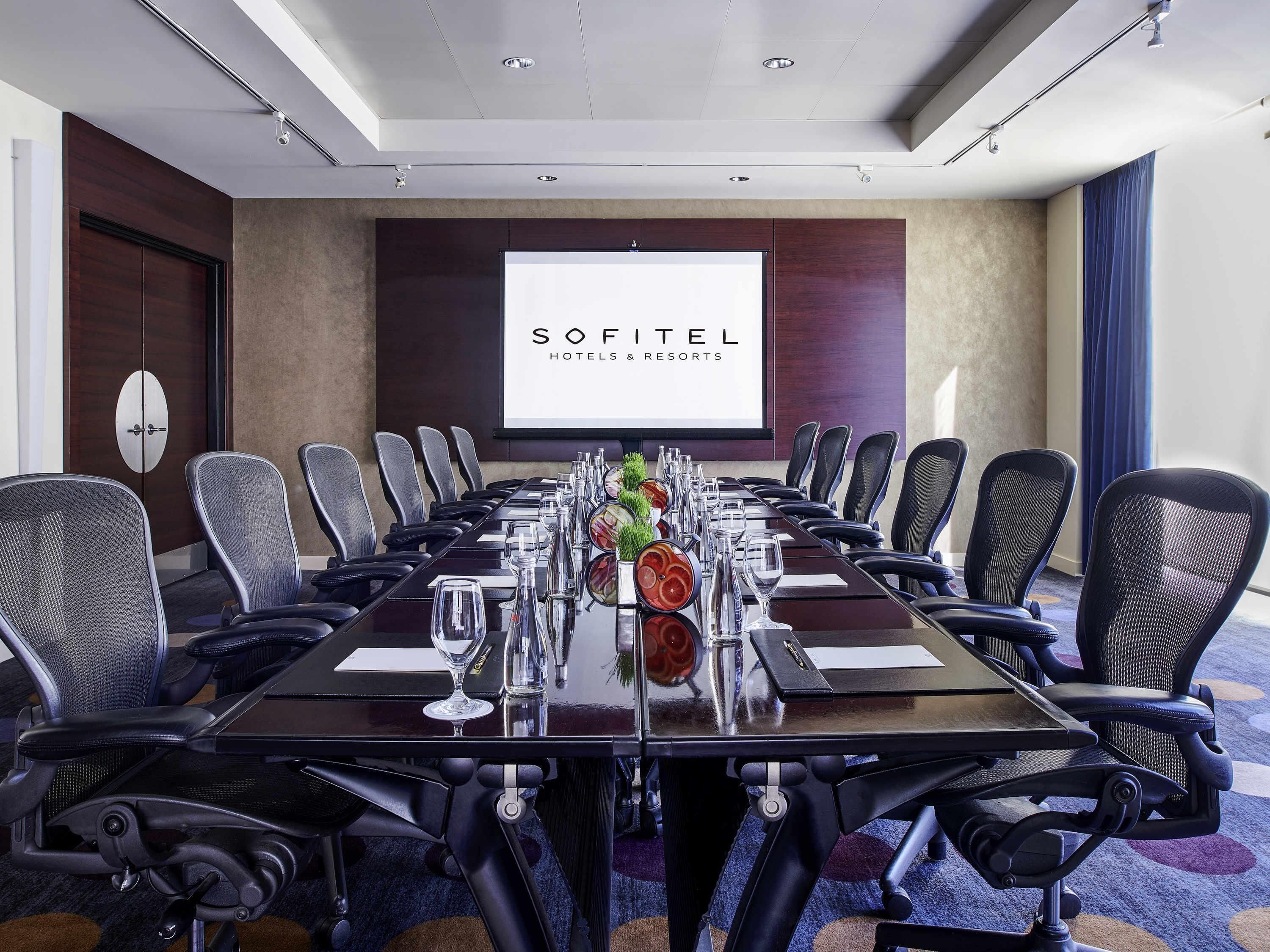 Uma sala de conferências com uma mesa comprida, cadeiras, um projetor e o logotipo Sofitel Hotels & Resorts.