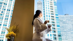 Une personne vêtue d’une robe blanche est assise près d’une fenêtre, d’où l’on peut admirer des immeubles modernes de grande hauteur.