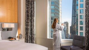 Une chambre d’hôtel avec un grand lit, une chaise, une fenêtre donnant sur la ville et une salle de bain équipée de toilettes et d’un lavabo.