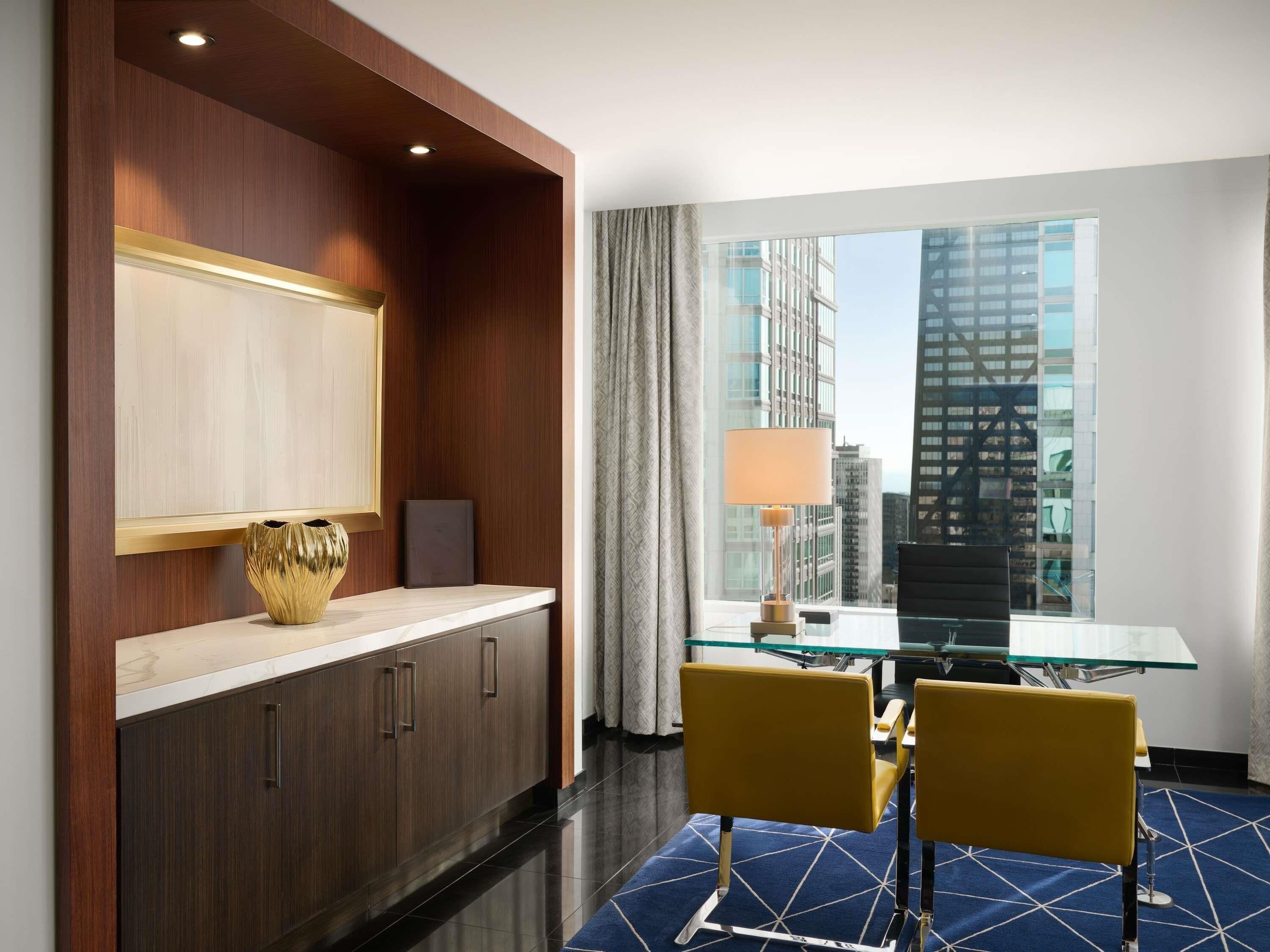 Habitación de hotel con zona de comedor, un amplio ventanal con vistas a la ciudad, escritorio con lámpara y una silla.