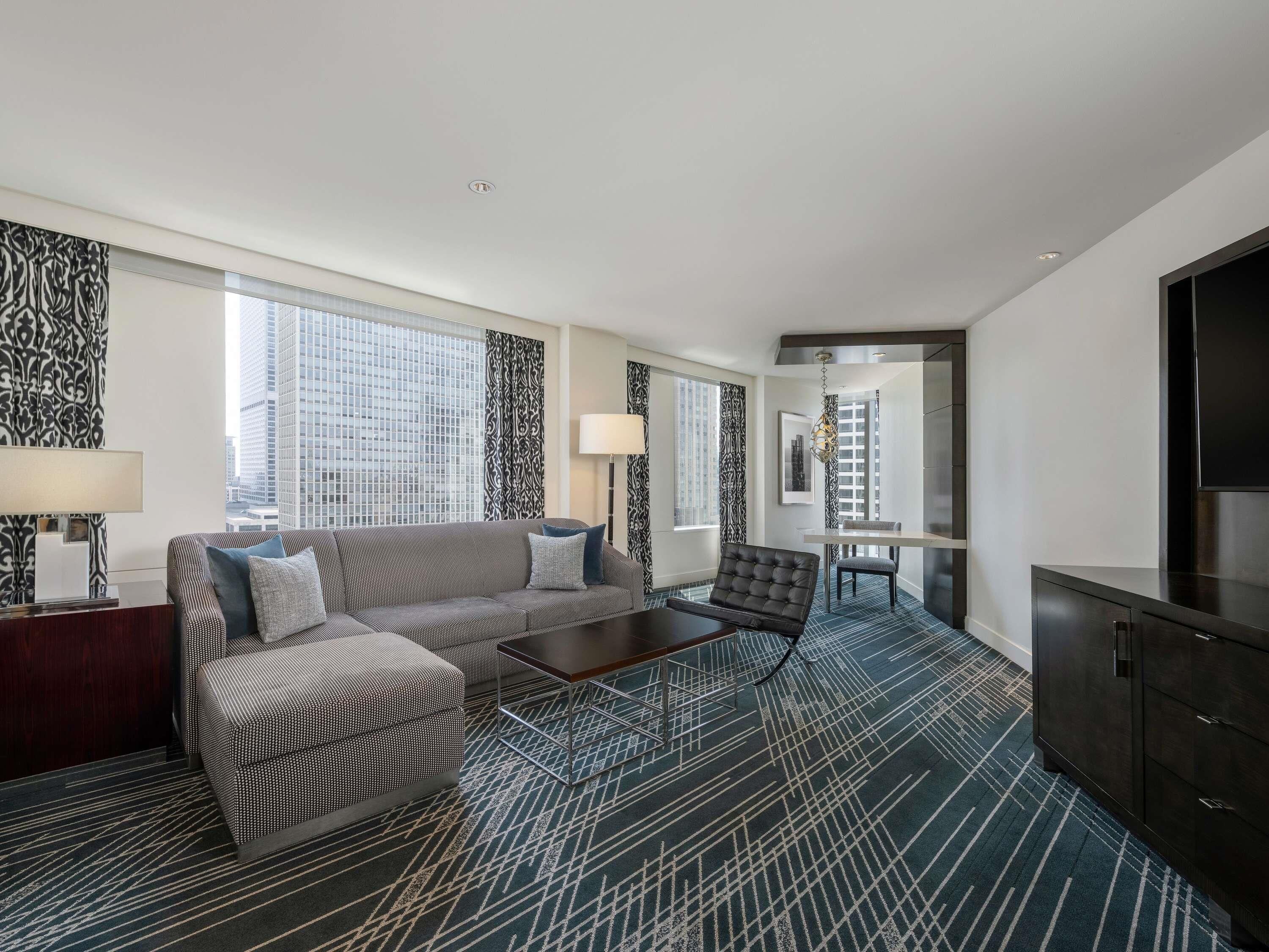 A modern hotel room with a sofa, ottoman, coffee table, and a TV.