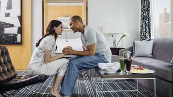 Un couple est assis sur un lit dans une chambre d’hôtel moderne avec vue sur la ville. On aperçoit une table à manger avec de la nourriture et des boissons, ainsi qu’un canapé.