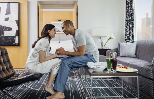 Un couple est assis sur un lit dans une chambre d’hôtel moderne avec vue sur la ville. On aperçoit une table à manger avec de la nourriture et des boissons, ainsi qu’un canapé.