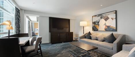 A modern hotel room with a sofa, armchairs, a coffee table, a TV, and a large window.
