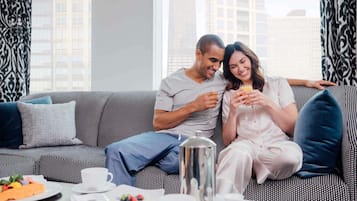 A couple sitting on a patterned sofa with a breakfast tray in front of them, including a coffee pot, cups, and a plate of fruit.