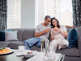 A couple sitting on a patterned sofa with a breakfast tray in front of them, including a coffee pot, cups, and a plate of fruit.