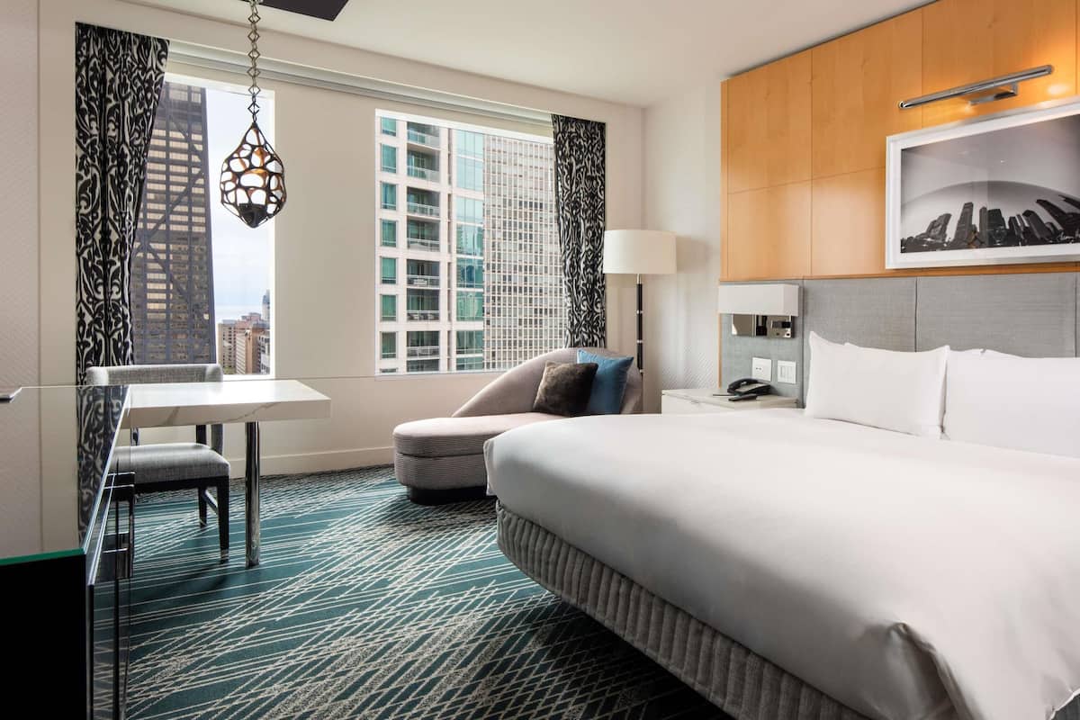 Habitación de hotel con cama, escritorio, silla, lámpara y vista a la ciudad.
