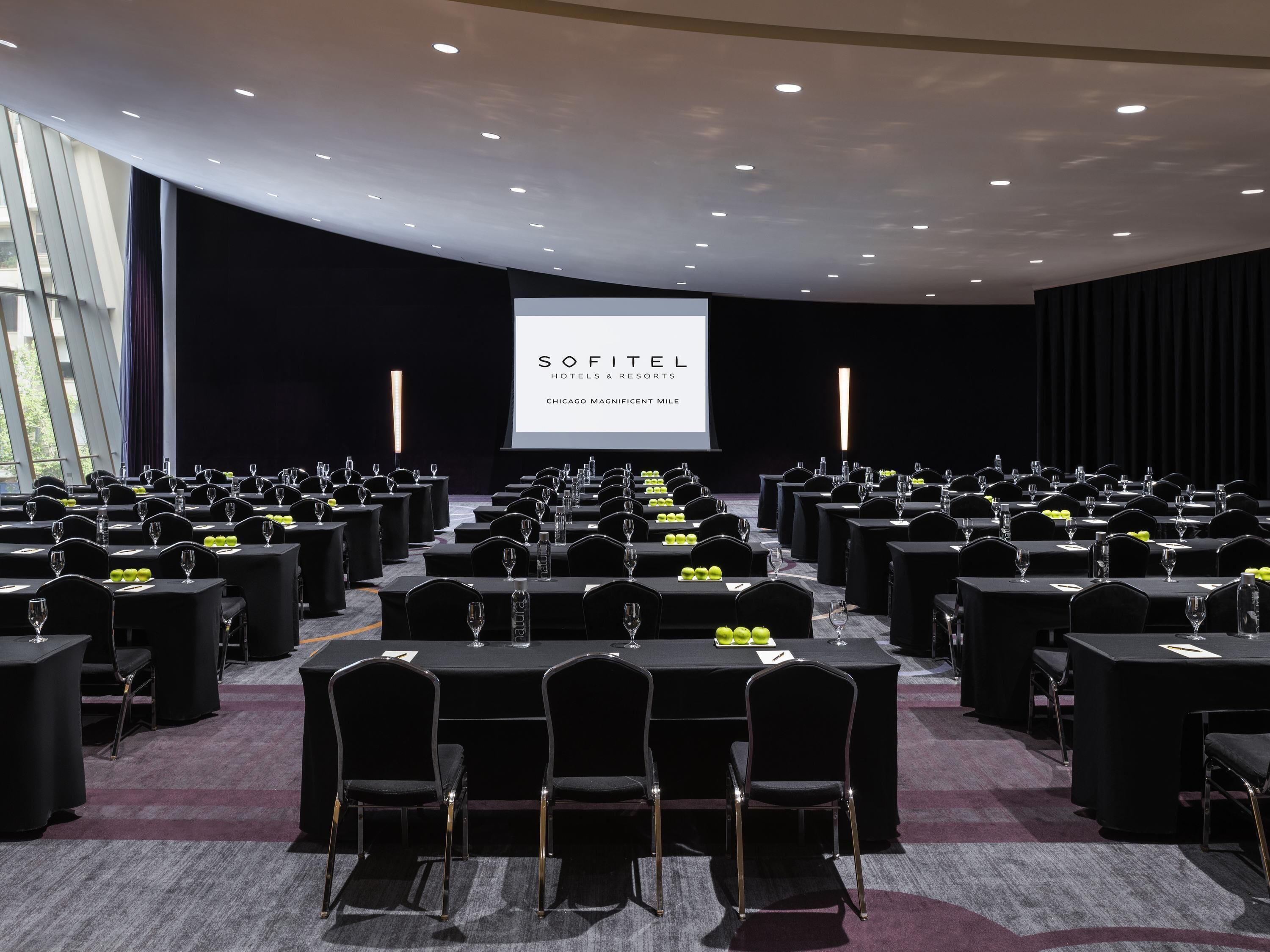 Une salle de conférence avec des rangées de tables et de chaises, un grand écran affichant le logo « SOFITEL HOTELS & RESORTS », et un intérieur moderne aux couleurs sombres.
