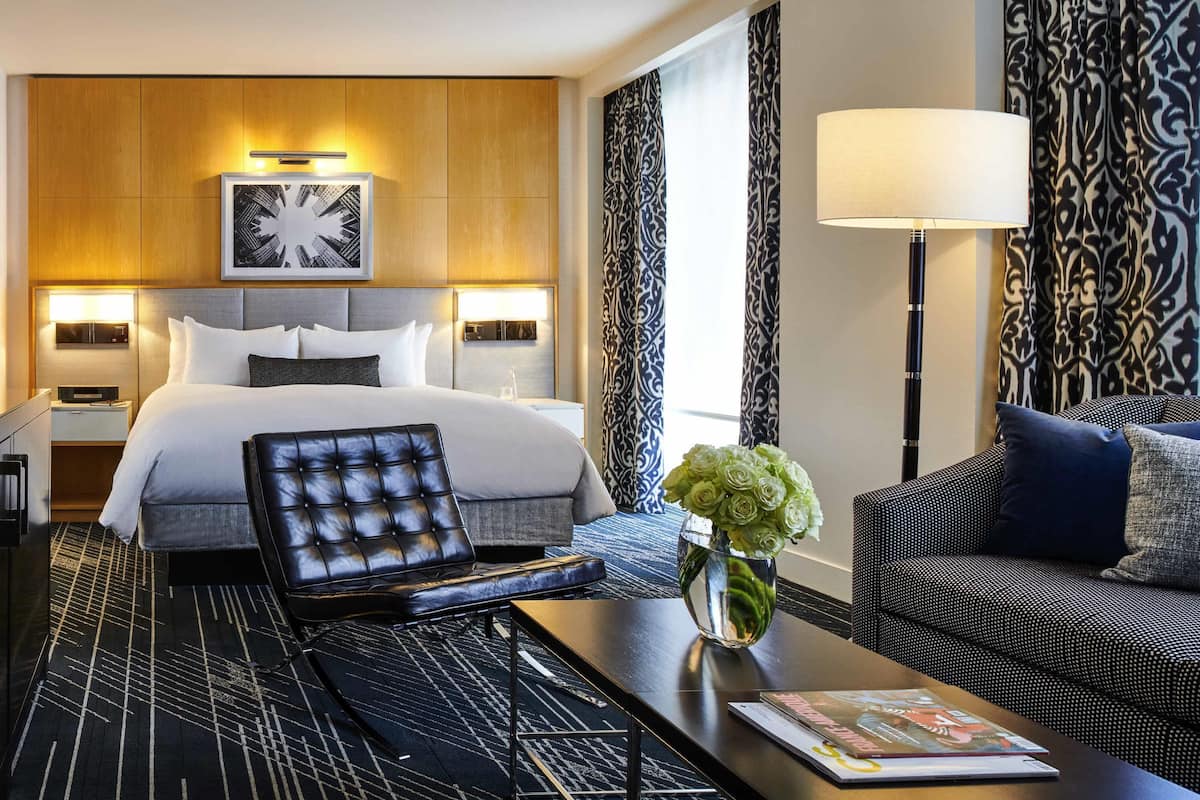 A hotel room with a bed, a sofa, a coffee table, a lamp, and a vase with flowers.