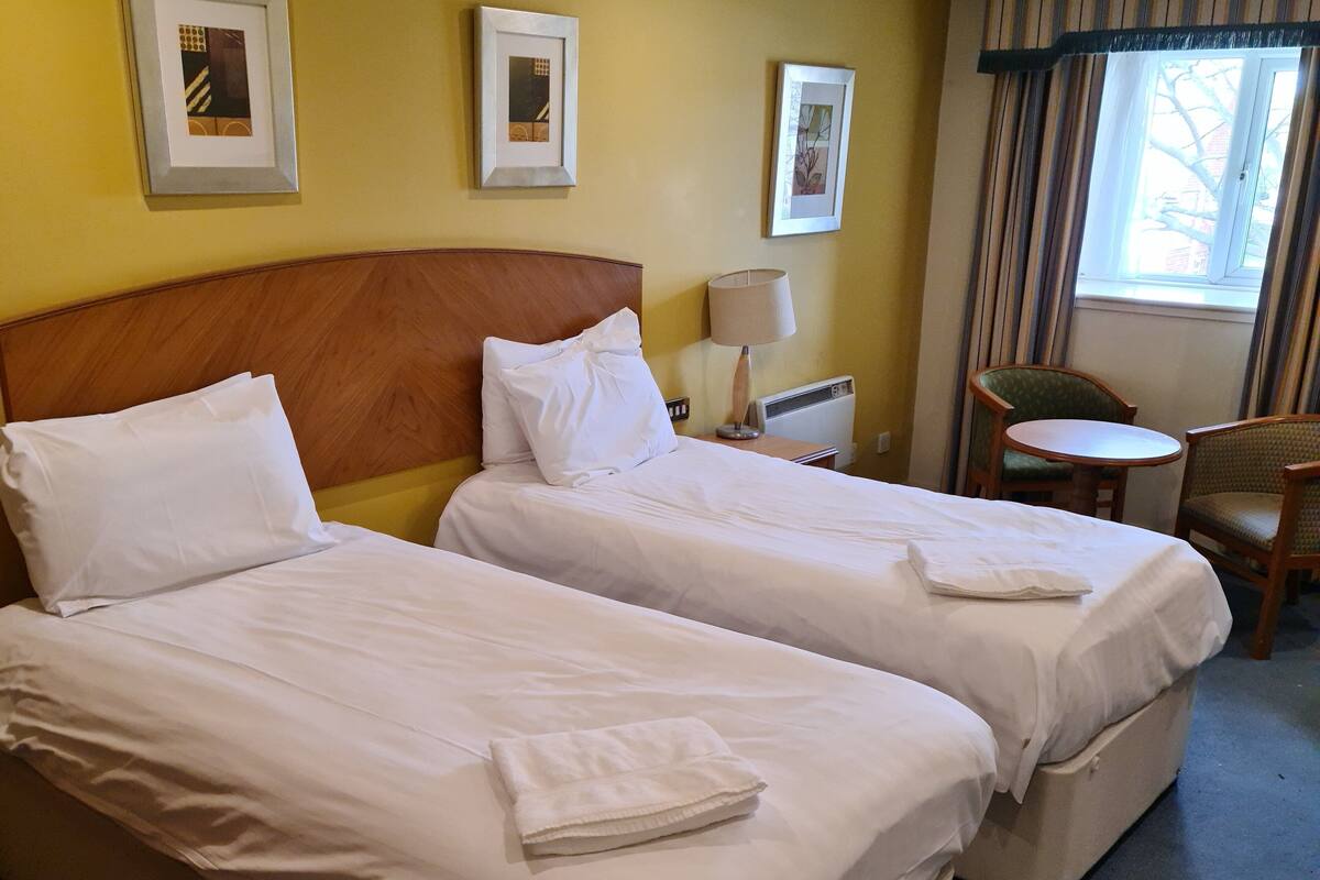A hotel room with two single beds, a small table, a chair, and a window with curtains.