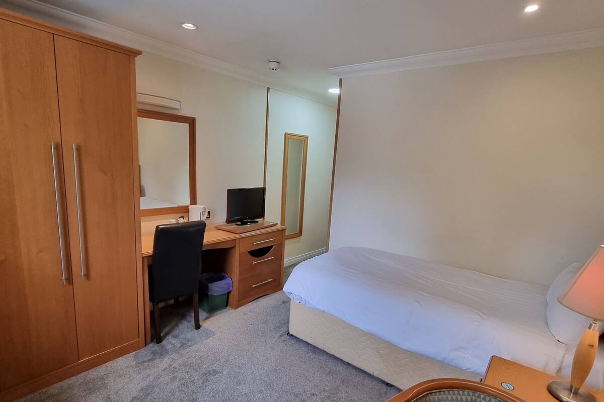 A hotel room with a bed, a desk with a computer, a chair, a wardrobe, and a bedside table with a lamp.