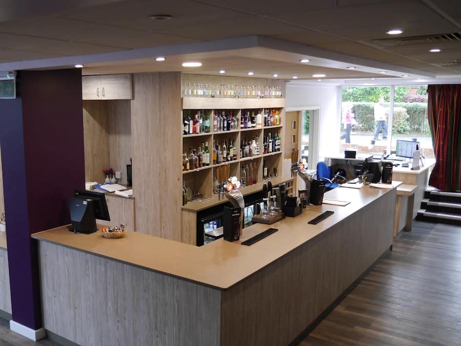 A bar area with a wooden counter, shelves with bottles, a coffee machine, and a computer monitor.