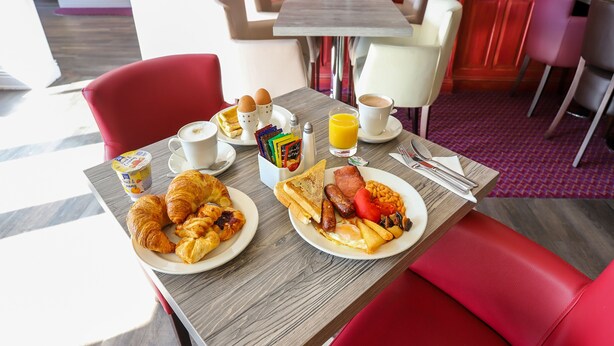 Una mesa de desayuno con croissants, huevos, tostadas, salchichas, frijoles y una taza de café.