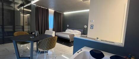 A modern hotel room with a large bed, a sitting area with a table and chairs, a bathtub, and a large mirror.