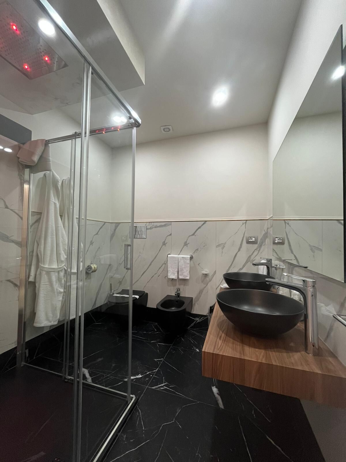 Un baño moderno con cabina de ducha de vidrio, un bidé negro, un tocador de madera con lavamanos negro y paredes de mármol.
