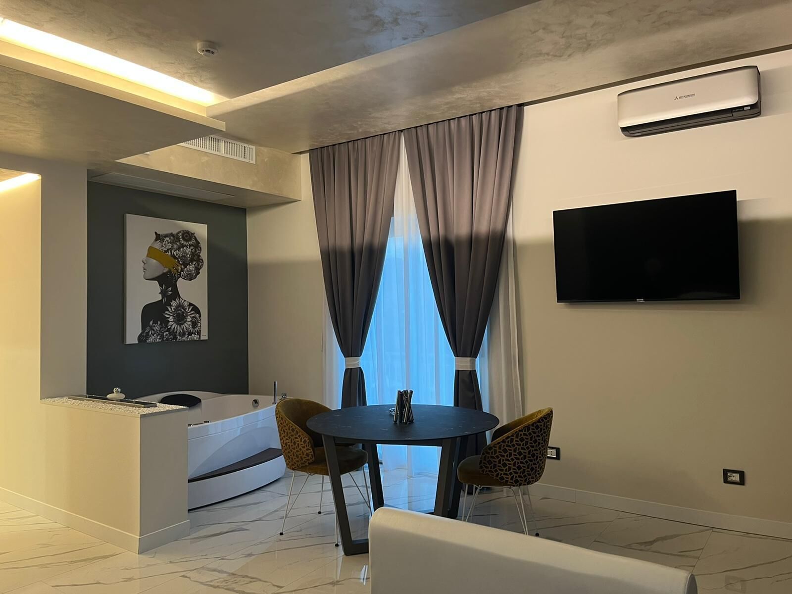 Une chambre d’hôtel moderne dotée d’une télévision à écran plat, d’un coin repas avec deux chaises, d’une petite table, d’un canapé et d’une grande fenêtre avec des rideaux.