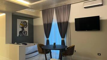 Une chambre d’hôtel moderne dotée d’une télévision à écran plat, d’un coin repas avec deux chaises, d’une petite table, d’un canapé et d’une grande fenêtre avec des rideaux.