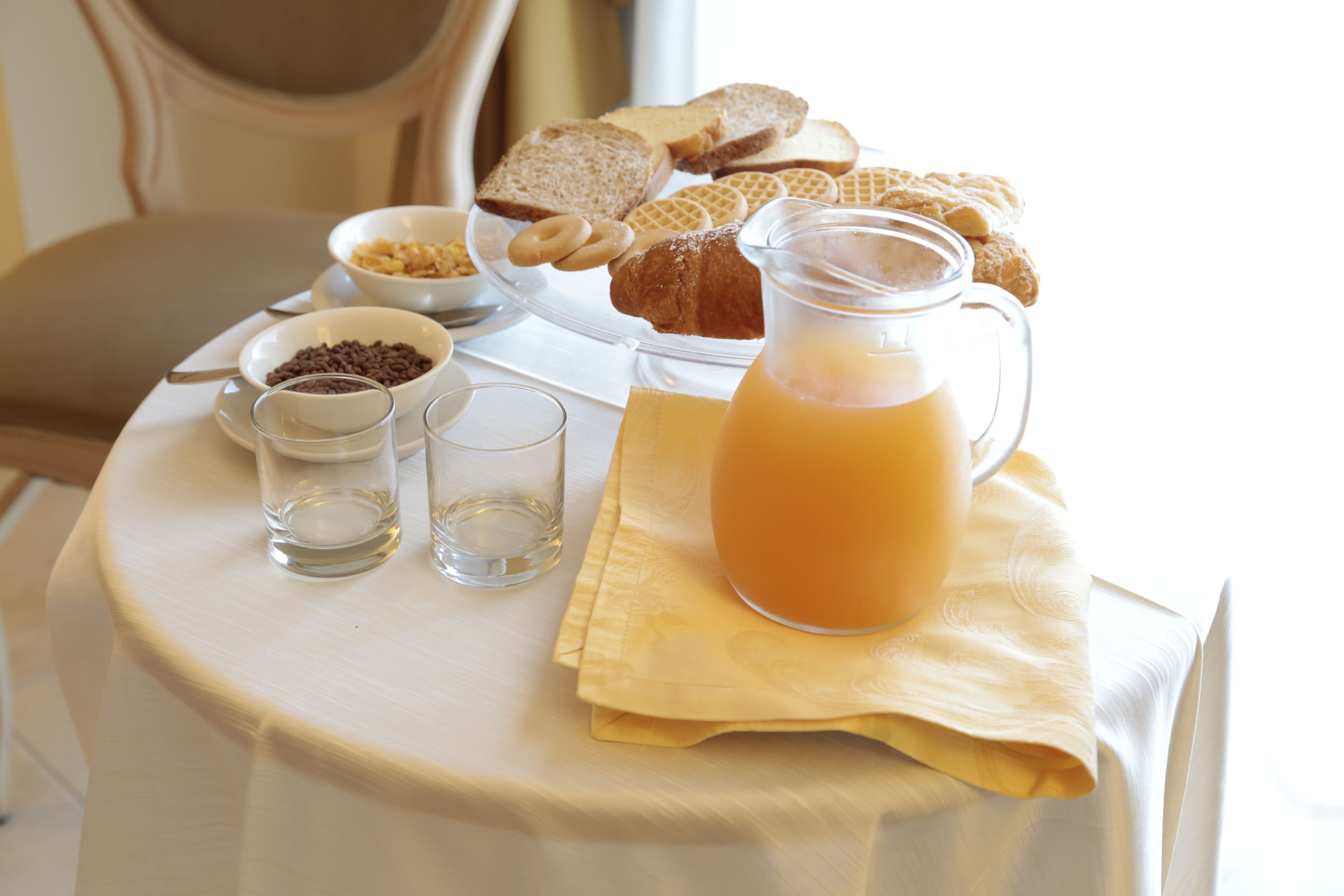 A breakfast table with bread, croissants, a pitcher of orange juice, and small bowls of cereal and coffee.