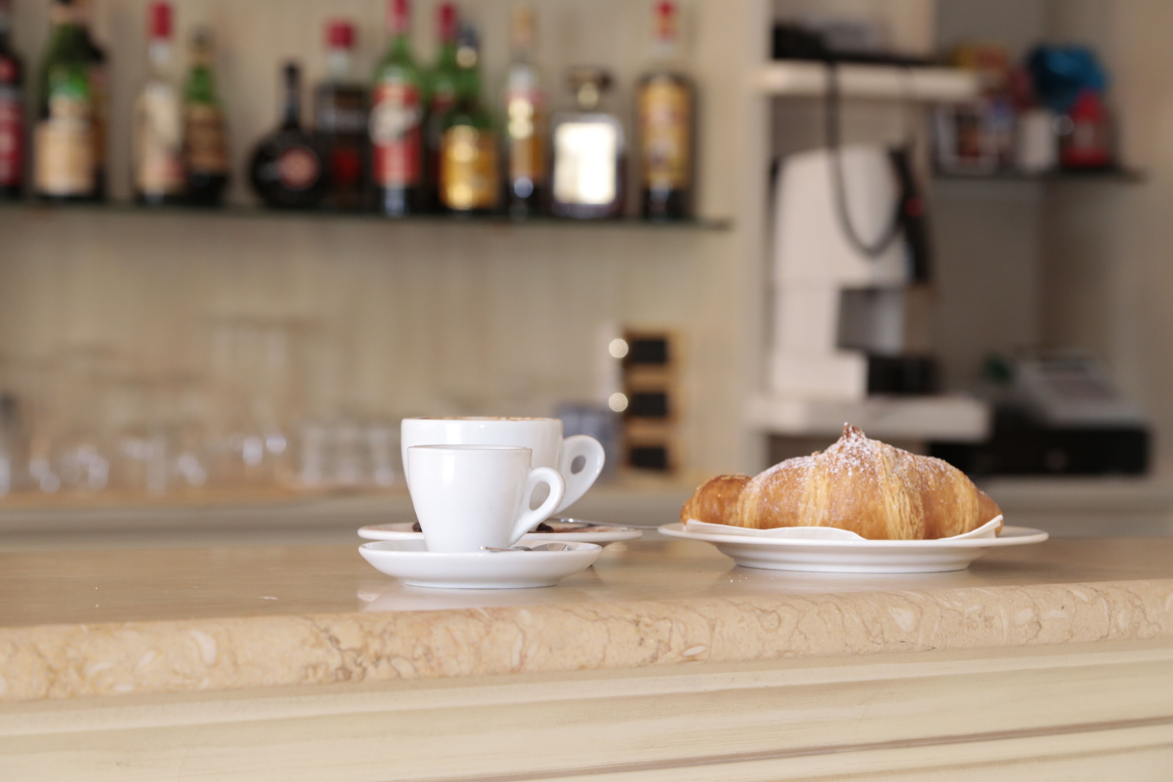 Una taza de café y un croissant sobre una encimera de mármol, con un fondo desenfocado de estantes con botellas.