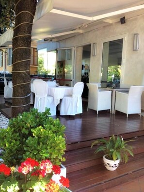 An outdoor dining area with white chairs and tables, surrounded by potted plants and a tree. - Hotel Mediterraneo (Pesaro)