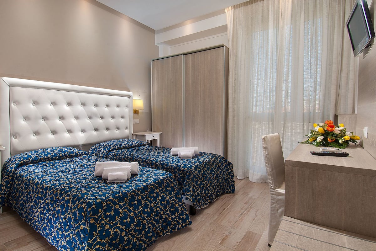 A hotel room with two beds, a desk, a TV, and a window with curtains.