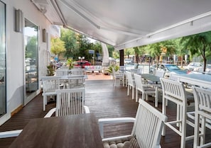 An outdoor patio area with tables and white chairs, a wooden deck, and a view of the street and parked cars. - Hotel Mediterraneo (Pesaro)