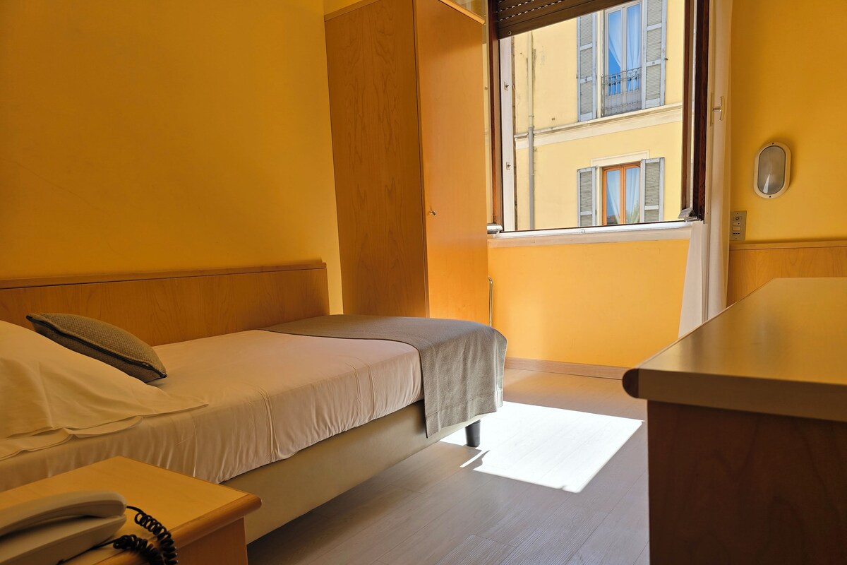 Una habitación con cama, mesita de noche y ventana con vista a edificios.