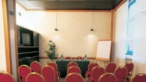 Ein Konferenzraum mit Reihen roter Stühle, einem Tisch mit grüner Tischdecke, einer Whiteboard und einer Pflanze.