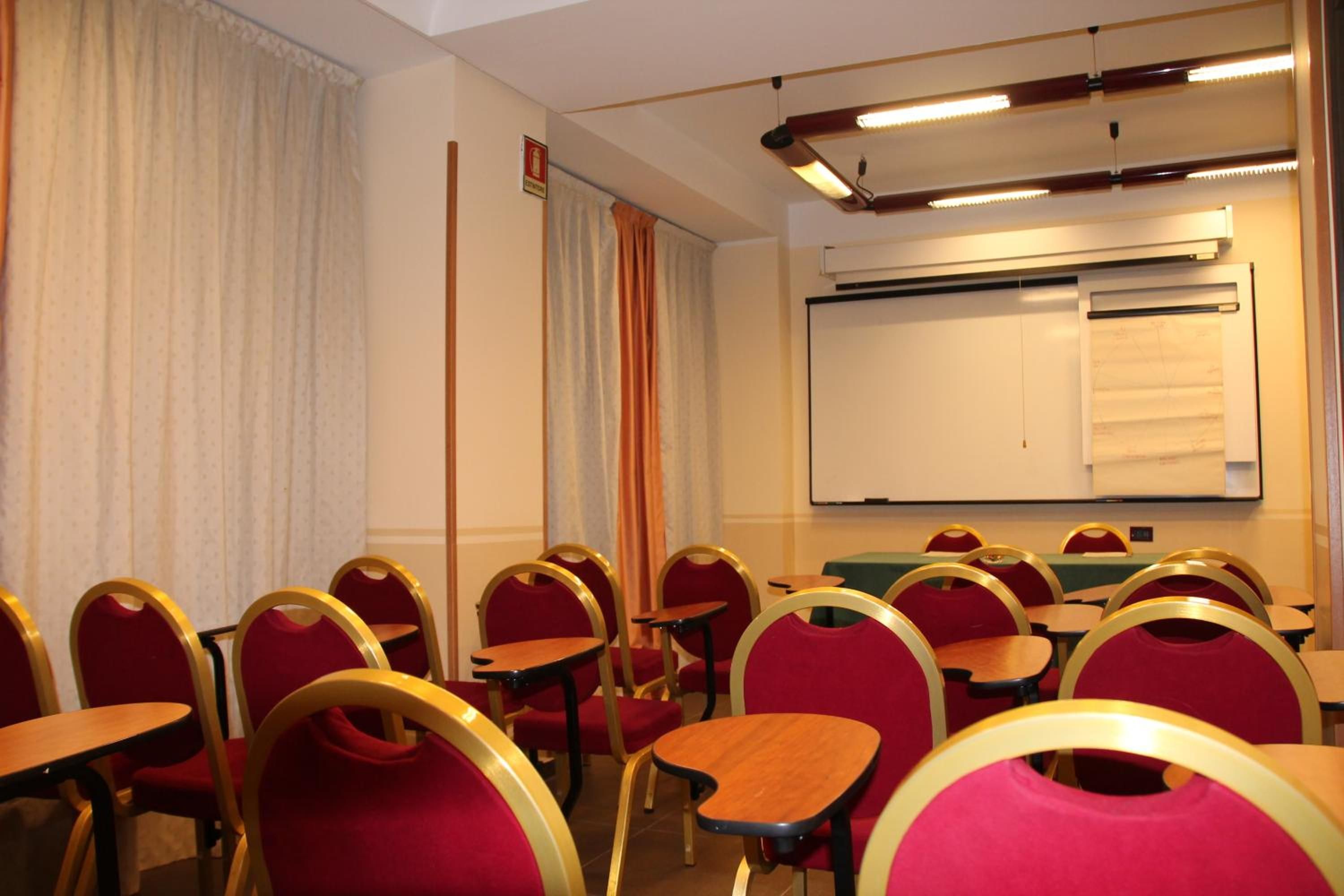Une salle de conférence avec des rangées de chaises rouges et des tables en bois, un tableau blanc et un appareil de climatisation fixé au plafond.
