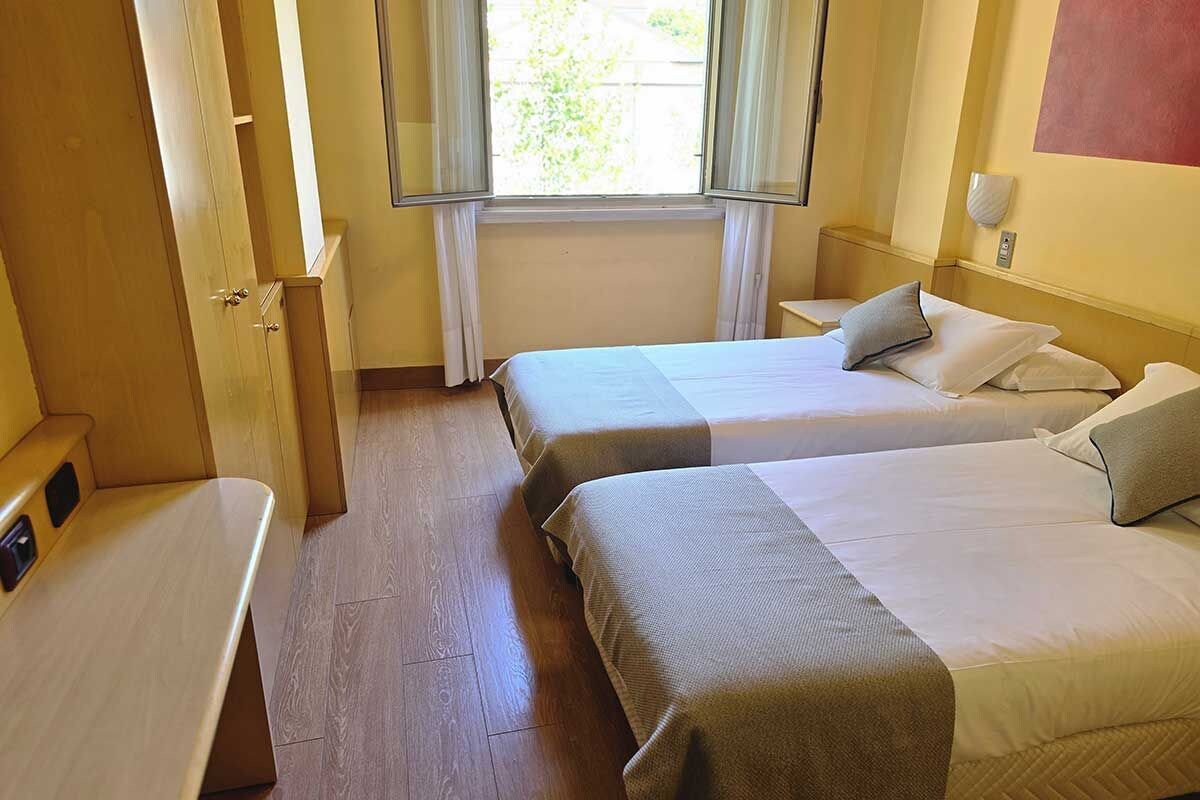 Habitación de hotel con dos camas individuales, un banco de madera, una ventana con cortinas y una lámpara fijada a la pared.